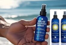 Solar Recovery Spray Reviews and Ingredients A person's hand holding a blue bottle of Solar Recovery Spray, with a blurred background of a sunny beach and calm ocean waves