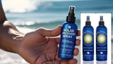 Solar Recovery Spray Reviews and Ingredients A person's hand holding a blue bottle of Solar Recovery Spray, with a blurred background of a sunny beach and calm ocean waves